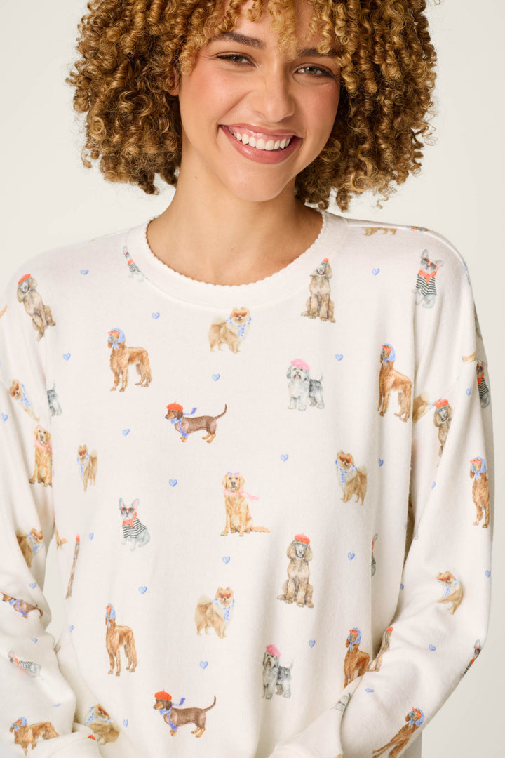 A smiling person with curly hair is wearing the P.J. Salvage "Meet Me in Pawris Dog Print Long Sleeve Top," a white sweater adorned with colorful illustrations of various dogs—some sporting hats or scarves—against a light background.