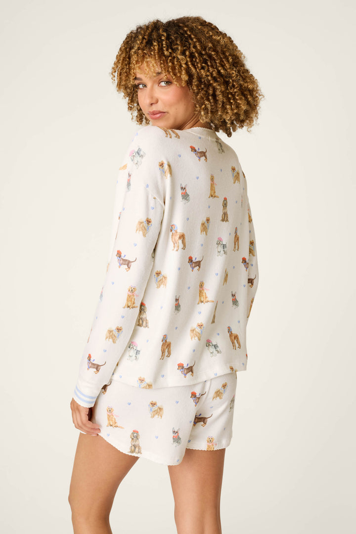 A woman with curly hair models the P.J. Salvage Meet Me in Pawris Dog Print Long Sleeve Top, paired with matching shorts. She stands with her back to the camera, glancing over her shoulder to highlight the playful dog print and relaxed fit against a simple, light background.