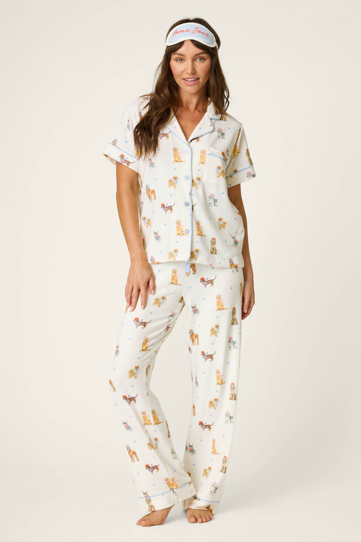 A woman stands barefoot against a plain background, smiling as she touches her hair, dressed in the P.J. Salvage Meet Me in Pawris Dog Print 3-Piece PJ Set with a matching sleep mask.