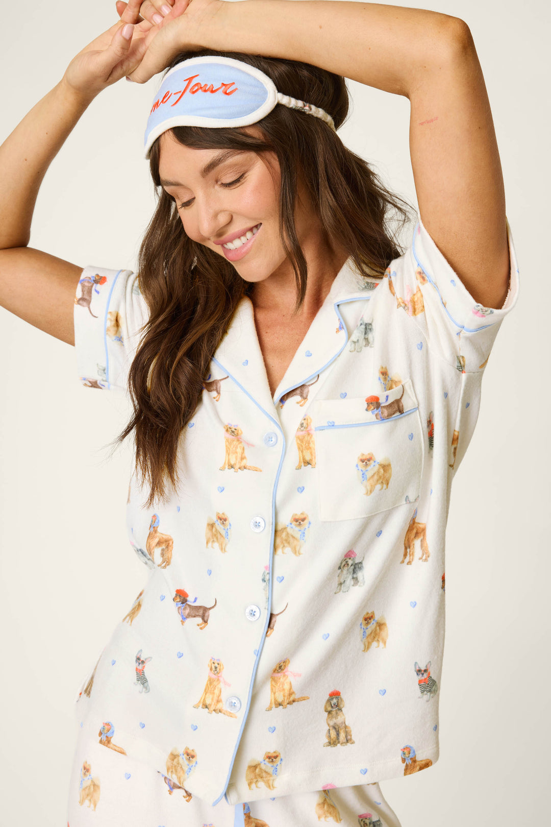 A woman smiles and stretches with arms raised, wearing the P.J. Salvage Meet Me in Pawris Dog Print 3-Piece PJ Set and a white sleep mask with red writing.