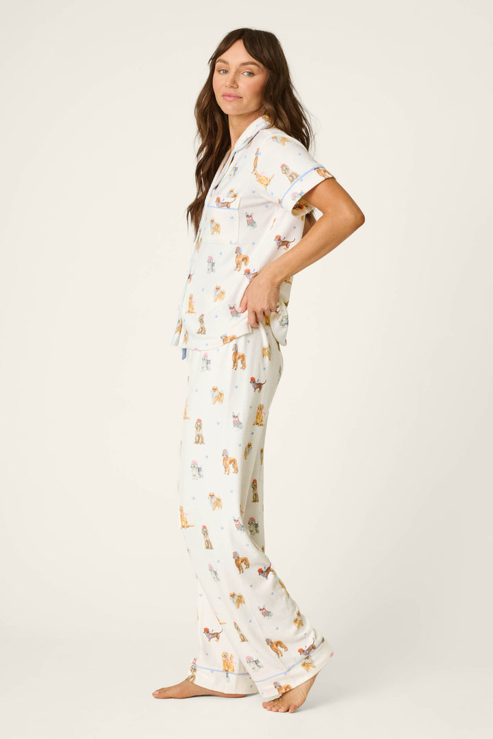 A woman stands sideways, barefoot, against a plain light background, wearing the P.J. Salvage Meet Me in Pawris Dog Print 3-Piece PJ Set—featuring a short-sleeved, collared pajama top and loose-fitting pants adorned with colorful dog prints.