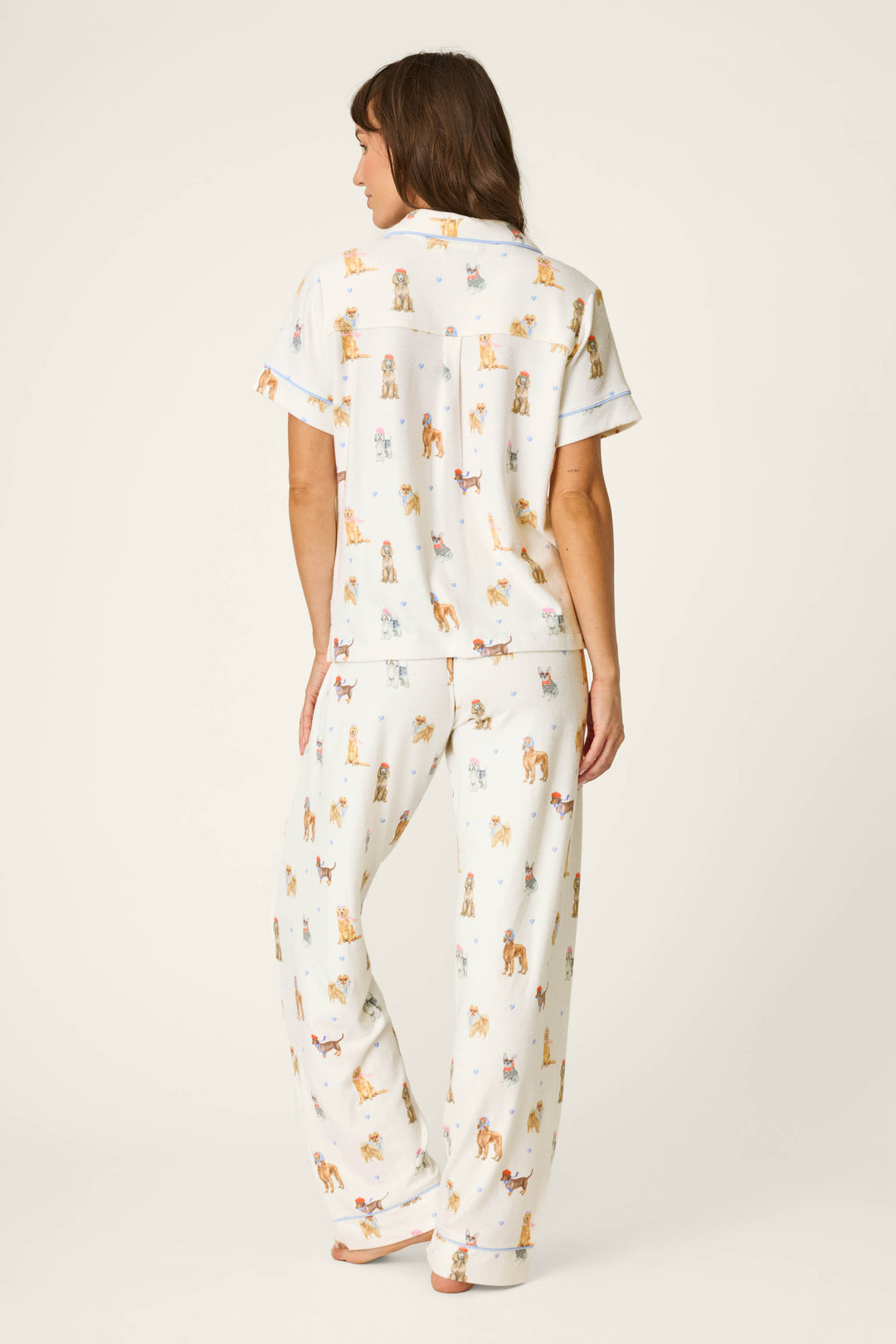A woman stands barefoot facing away, wearing the P.J. Salvage Meet Me in Pawris Dog Print 3-Piece PJ Set. The set features white pajamas with a colorful dog print, including a short-sleeve top and long pants, and she is posed against a plain, light background.