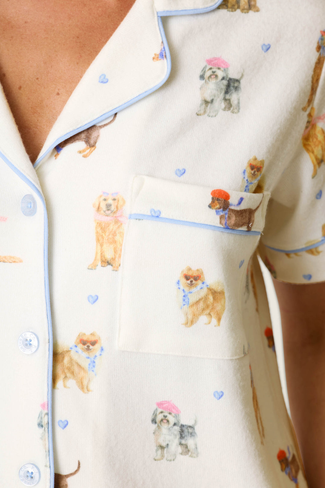 Close-up of the P.J. Salvage Meet Me in Pawris Dog Print 3-Piece PJ Set pajama top, featuring a cream-colored background with blue trim, patterned with playful illustrations of dogs in hats and scarves and accented by tiny blue hearts.