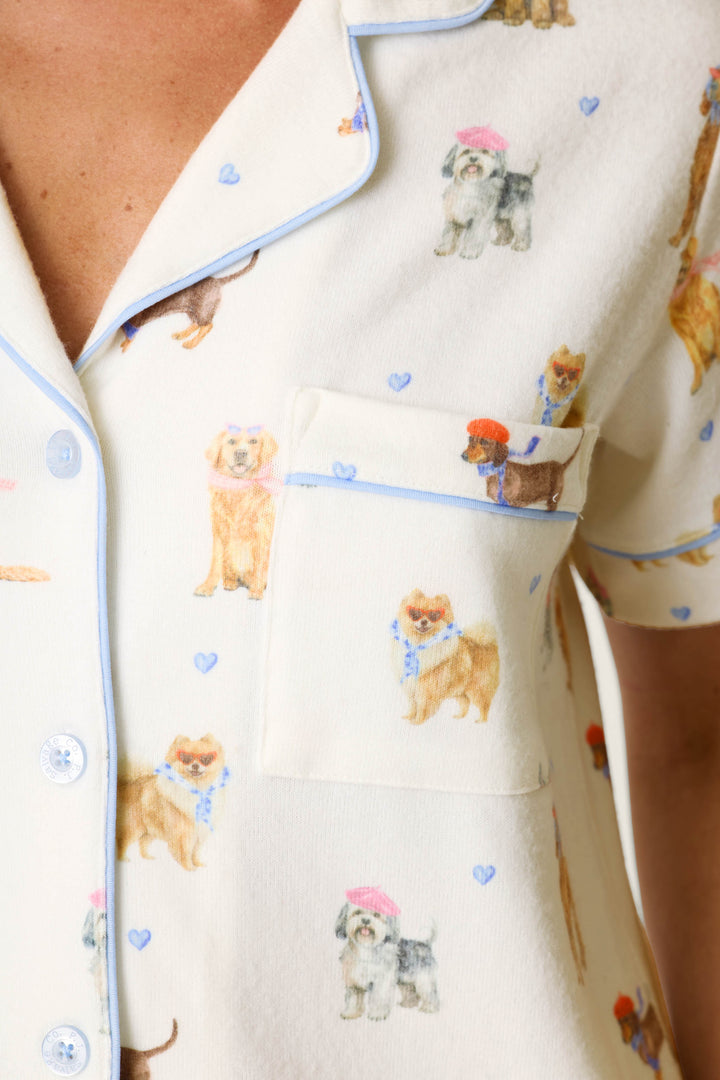 Close-up of the P.J. Salvage Meet Me in Pawris Dog Print 3-Piece PJ Set pajama top, featuring a cream-colored background with blue trim, patterned with playful illustrations of dogs in hats and scarves and accented by tiny blue hearts.