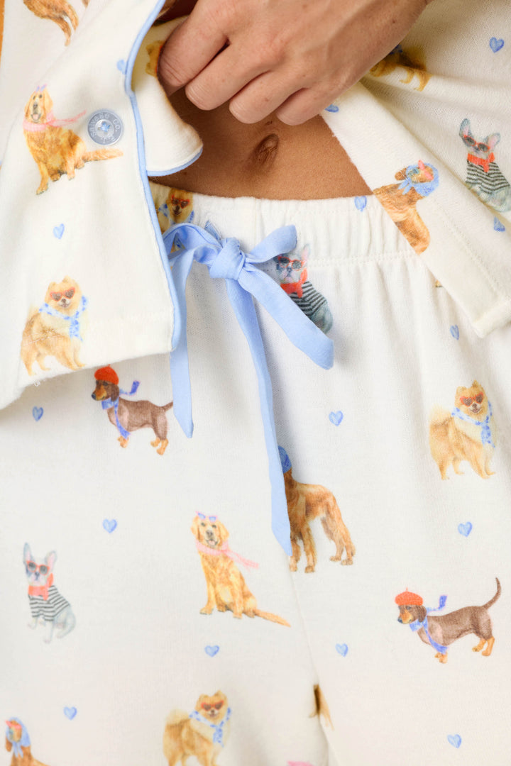 Close-up of the Meet Me in Pawris Dog Print 3-Piece PJ Set by P.J. Salvage, featuring pajama pants with a blue ribbon drawstring and a playful dog pattern. Various dog breeds are illustrated wearing hats and scarves, while small blue hearts are scattered across the fabric.