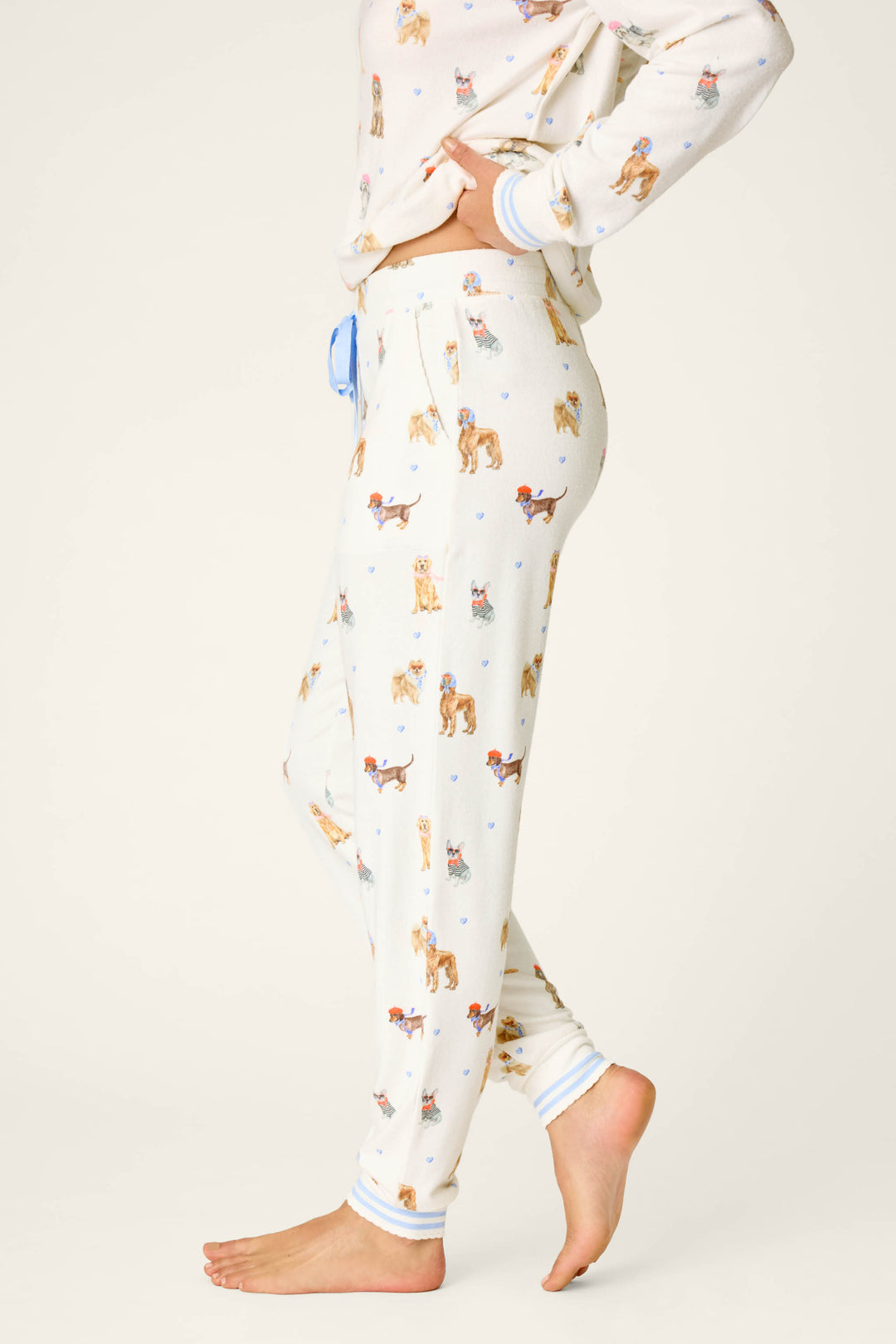 A person stands barefoot in side profile, showcasing the lower half and torso while wearing the P.J. Salvage Meet Me in Pawris Dog Print Jogger Pajama Pants, featuring a cream base and colorful dog illustrations, set against a plain background.
