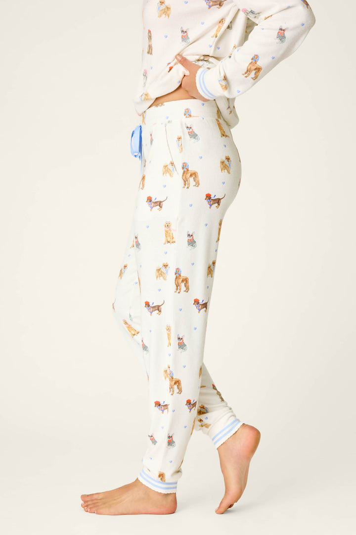 A person stands barefoot in side profile, showcasing the lower half and torso while wearing the P.J. Salvage Meet Me in Pawris Dog Print Jogger Pajama Pants, featuring a cream base and colorful dog illustrations, set against a plain background.