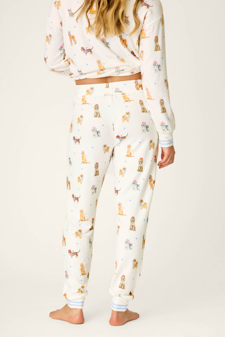 A woman stands barefoot with her back to the camera, wearing P.J. Salvage's "Meet Me in Pawris Dog Print Jogger Pajama Pants" featuring colorful dog illustrations. Only her back and lower legs are visible against a plain, light-toned background.