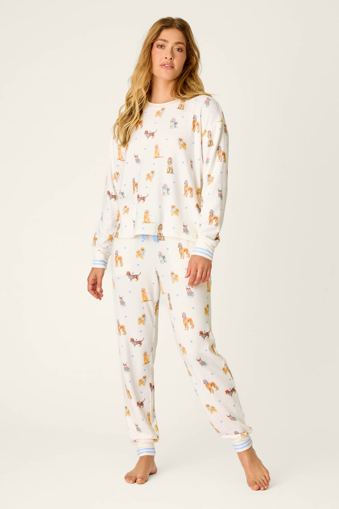 A woman with long hair models the Meet Me in Pawris Dog Print Long Sleeve PJ Set by P.J. Salvage—cozy white pajamas adorned with a colorful dog print. She stands barefoot against a plain, light background, facing forward with a relaxed expression and one hand by her side.