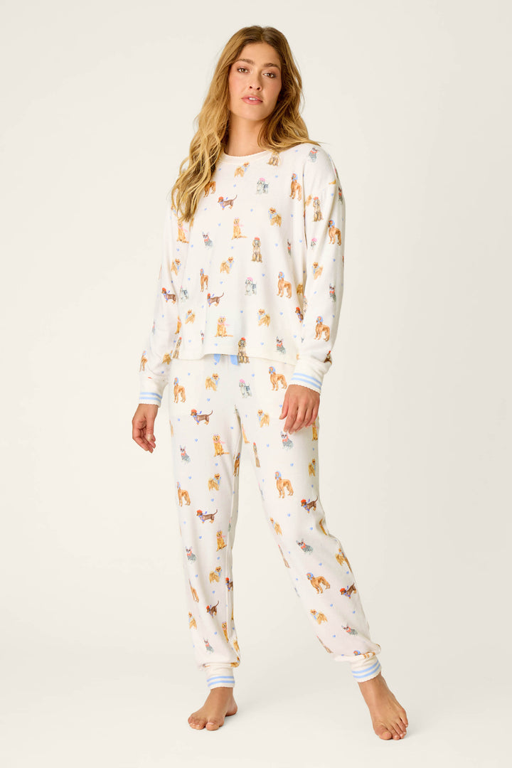 A woman with long hair models the Meet Me in Pawris Dog Print Long Sleeve PJ Set by P.J. Salvage—cozy white pajamas adorned with a colorful dog print. She stands barefoot against a plain, light background, facing forward with a relaxed expression and one hand by her side.
