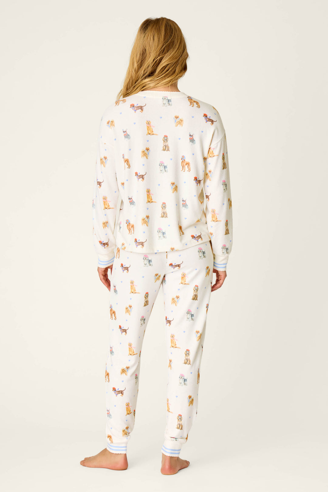 A person stands barefoot facing away, wearing the P.J. Salvage Meet Me in Pawris Dog Print Long Sleeve PJ Set, which features a white background with colorful dog prints. The pajama set includes long sleeves and pants, and is shown against a plain light-colored backdrop.