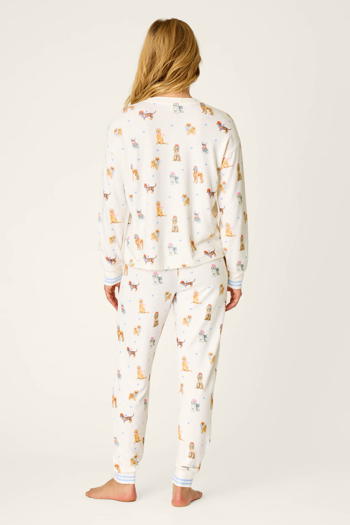 A person stands barefoot facing away, wearing the P.J. Salvage Meet Me in Pawris Dog Print Long Sleeve PJ Set, which features a white background with colorful dog prints. The pajama set includes long sleeves and pants, and is shown against a plain light-colored backdrop.