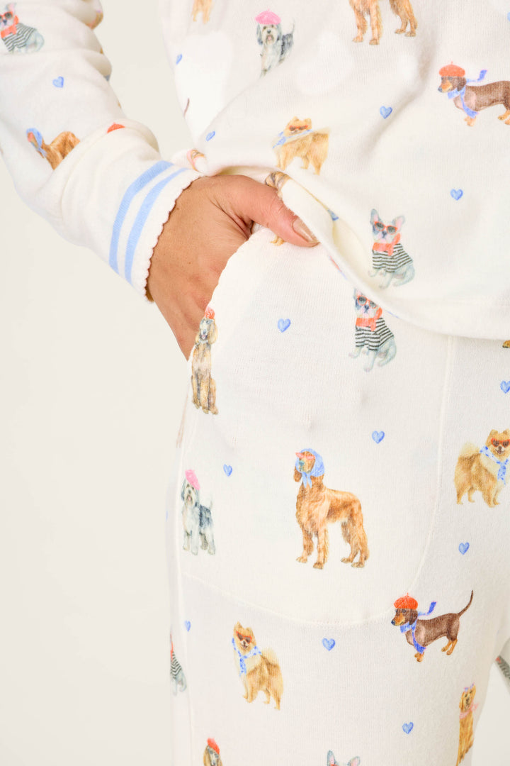 A model wears the P.J. Salvage "Meet Me in Pawris" Dog Print Long Sleeve PJ Set featuring a white background with colorful dog illustrations and small blue hearts, posing with one hand in the pocket. Only the torso and upper legs are shown against a plain backdrop.