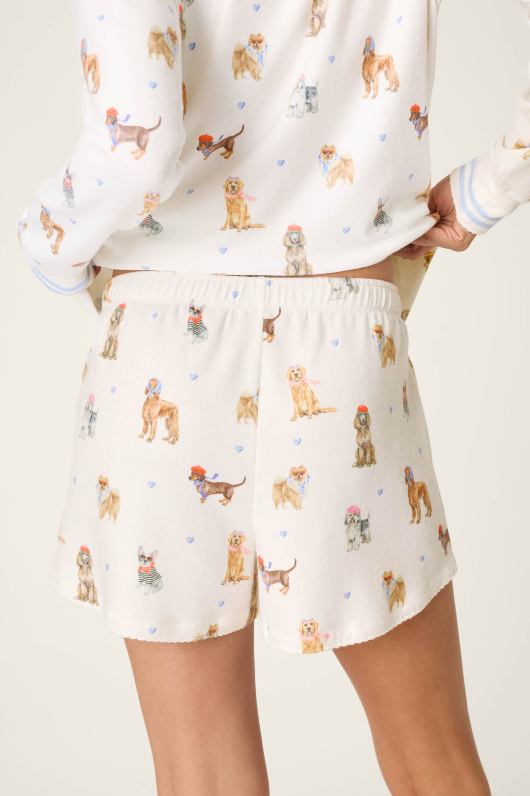 A person is seen from the back wearing the P.J. Salvage Meet Me in Pawris Dog Print Pajama Shorts, featuring colorful dog illustrations and small blue hearts; the matching pajama top is partially visible.