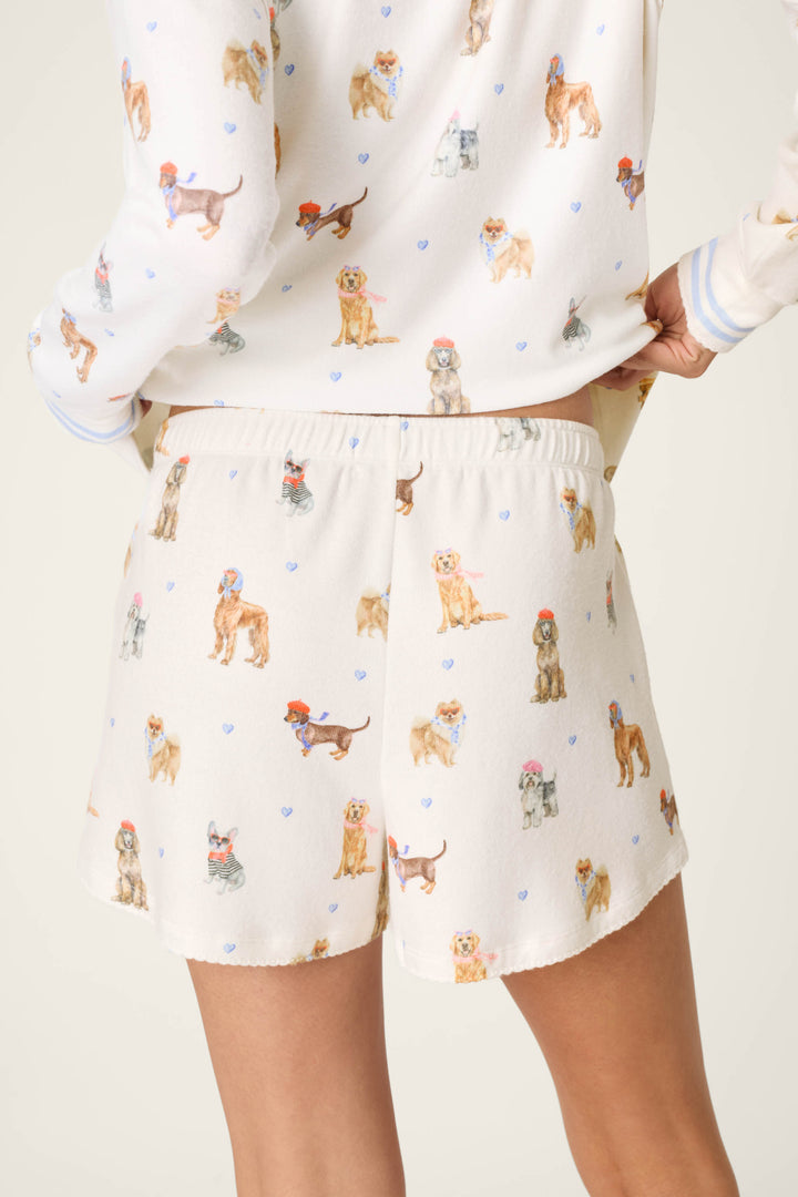 A person is seen from the back wearing the P.J. Salvage Meet Me in Pawris Dog Print Pajama Shorts, featuring colorful dog illustrations and small blue hearts; the matching pajama top is partially visible.