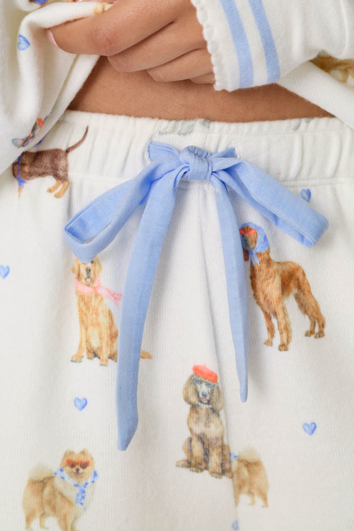 Close-up of the P.J. Salvage Meet Me in Pawris Dog Print Pajama Shorts, featuring a blue bow at the waistband and an illustrated dog pattern with hats, scarves, and small blue hearts. The wearer’s hand rests near the waistband.