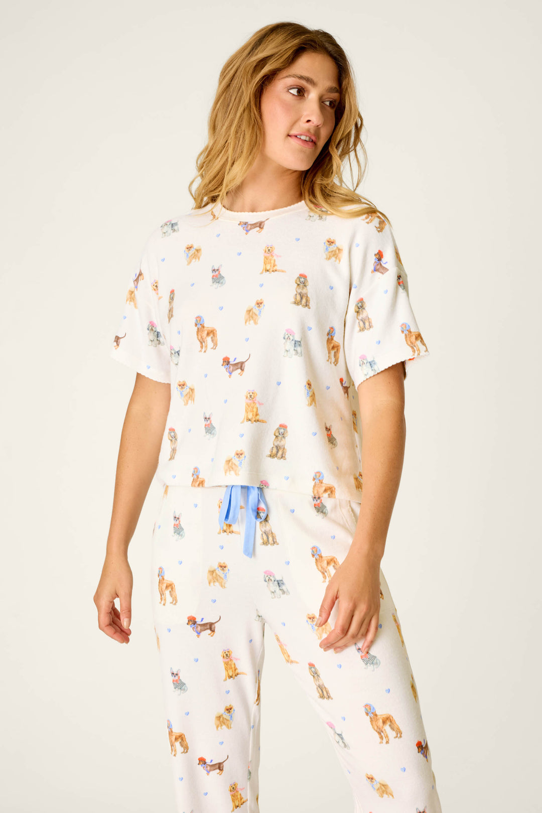 A woman stands against a plain background, looking slightly to her left with a relaxed expression, wearing the P.J. Salvage "Meet Me in Pawris Dog Print" short sleeve top featuring a colorful dog print and blue drawstring.
