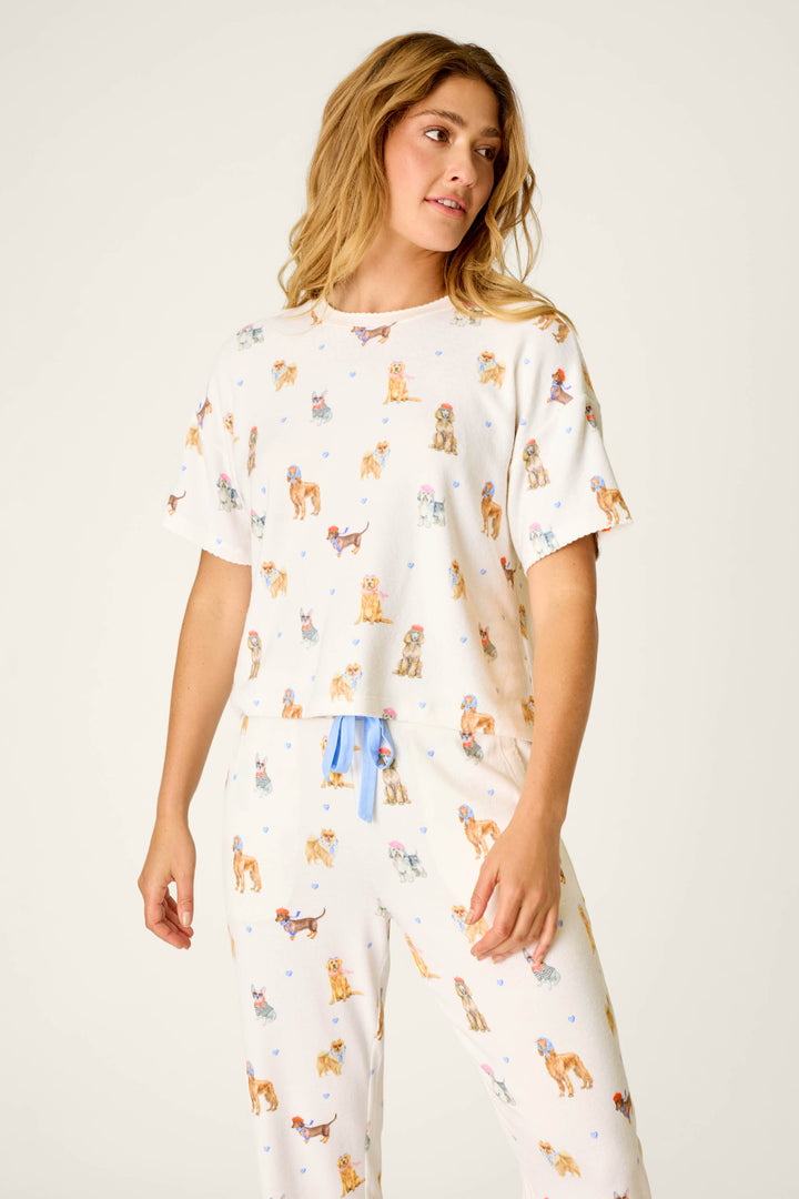 A woman stands against a plain background, looking slightly to her left with a relaxed expression, wearing the P.J. Salvage "Meet Me in Pawris Dog Print" short sleeve top featuring a colorful dog print and blue drawstring.