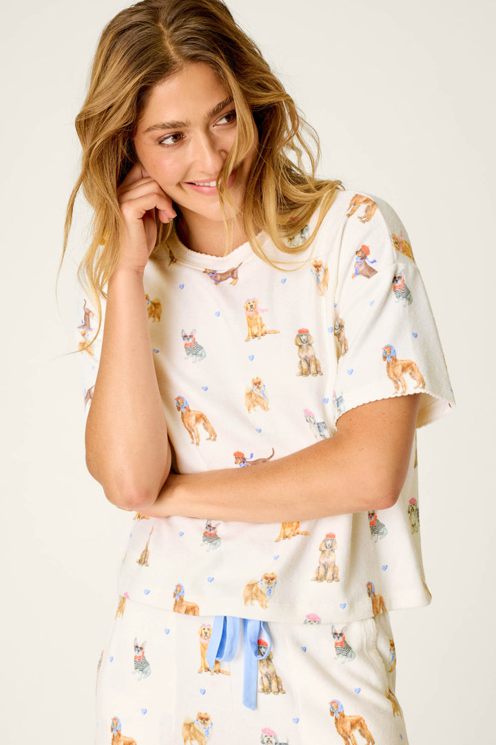 A smiling woman with long blonde hair wears the P.J. Salvage "Meet Me in Pawris" Dog Print Short Sleeve Top—cream-colored pajamas decorated with cartoon dogs and blue hearts—while standing against a plain background.