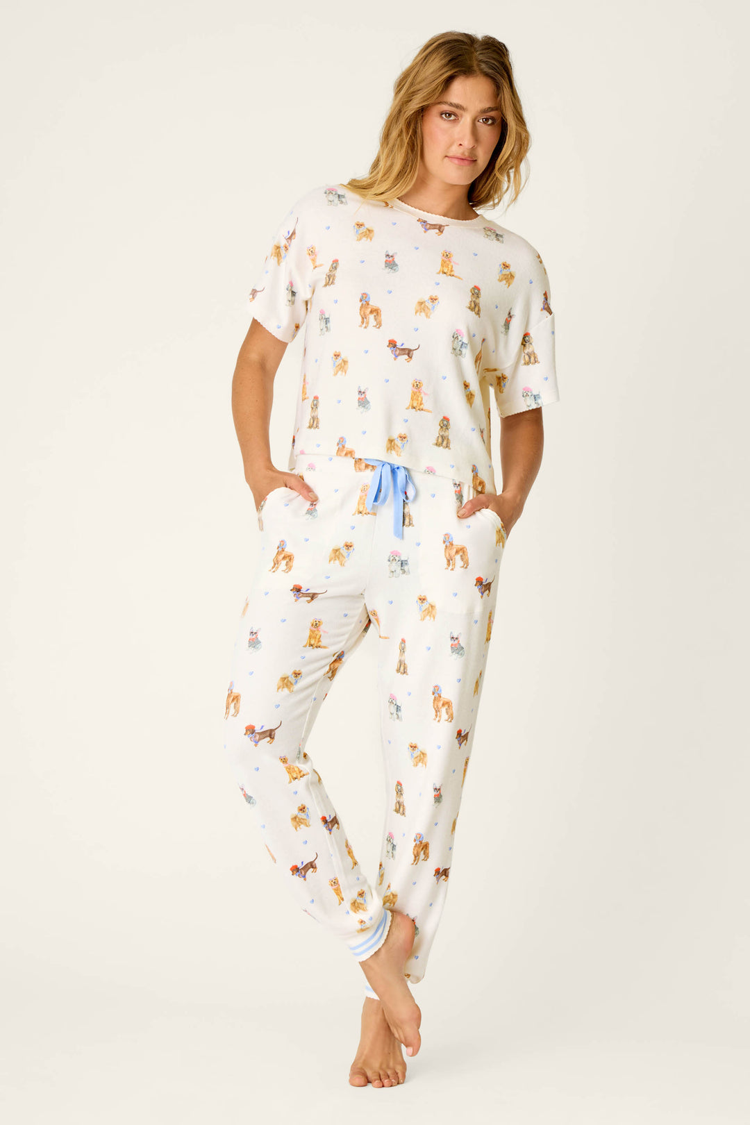 A woman stands barefoot with light brown wavy hair and a relaxed expression, wearing the P.J. Salvage "Meet Me in Pawris" Dog Print Short Sleeve Top paired with matching white pajamas featuring a colorful dog print, a blue drawstring, and side pockets.