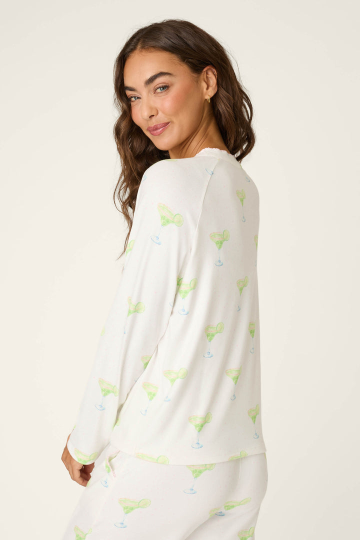 A woman with long brown hair, wearing the P.J. Salvage Margarita Mama Spicy Sips Long Sleeve Top in a white jersey with green margarita prints, stands facing slightly away and smiles over her shoulder.