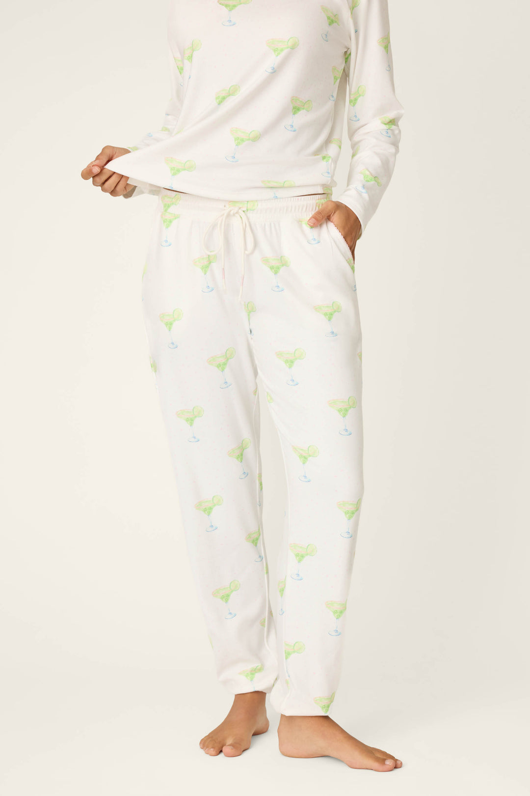 A person wears P.J. Salvage Margarita Mama Spicy Sips Banded Pants featuring a green and blue margarita print, standing barefoot with one hand in a pocket and the other holding the shirt hem against a plain background.