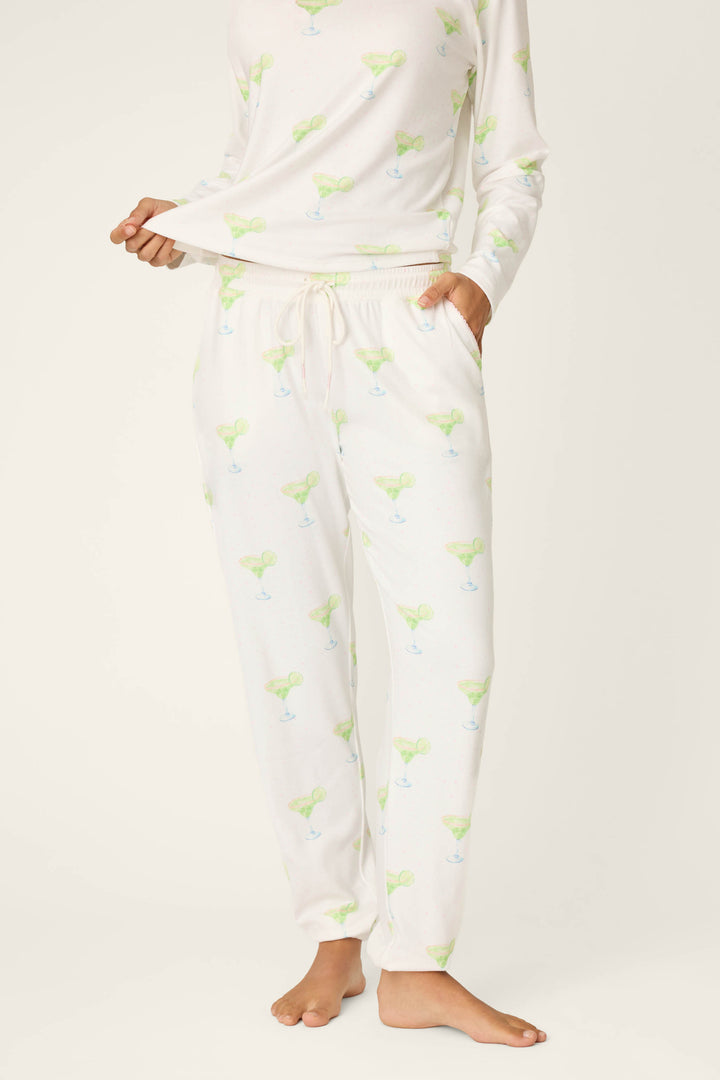 A person wears P.J. Salvage Margarita Mama Spicy Sips Banded Pants featuring a green and blue margarita print, standing barefoot with one hand in a pocket and the other holding the shirt hem against a plain background.