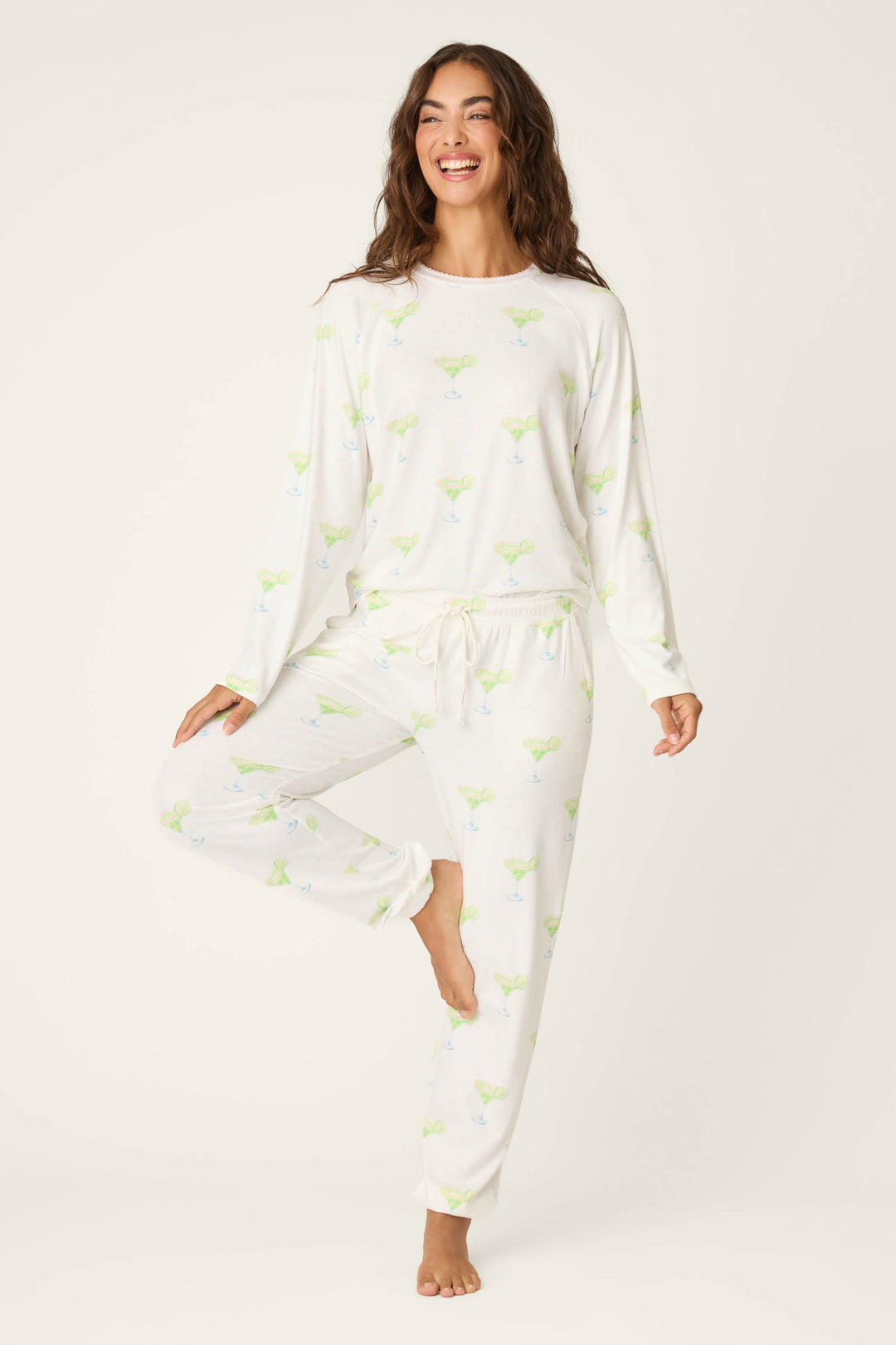 A woman smiles in a relaxed pose, wearing P.J. Salvage’s Margarita Mama Spicy Sips Banded Pants with a playful margarita print, set against a plain light background.