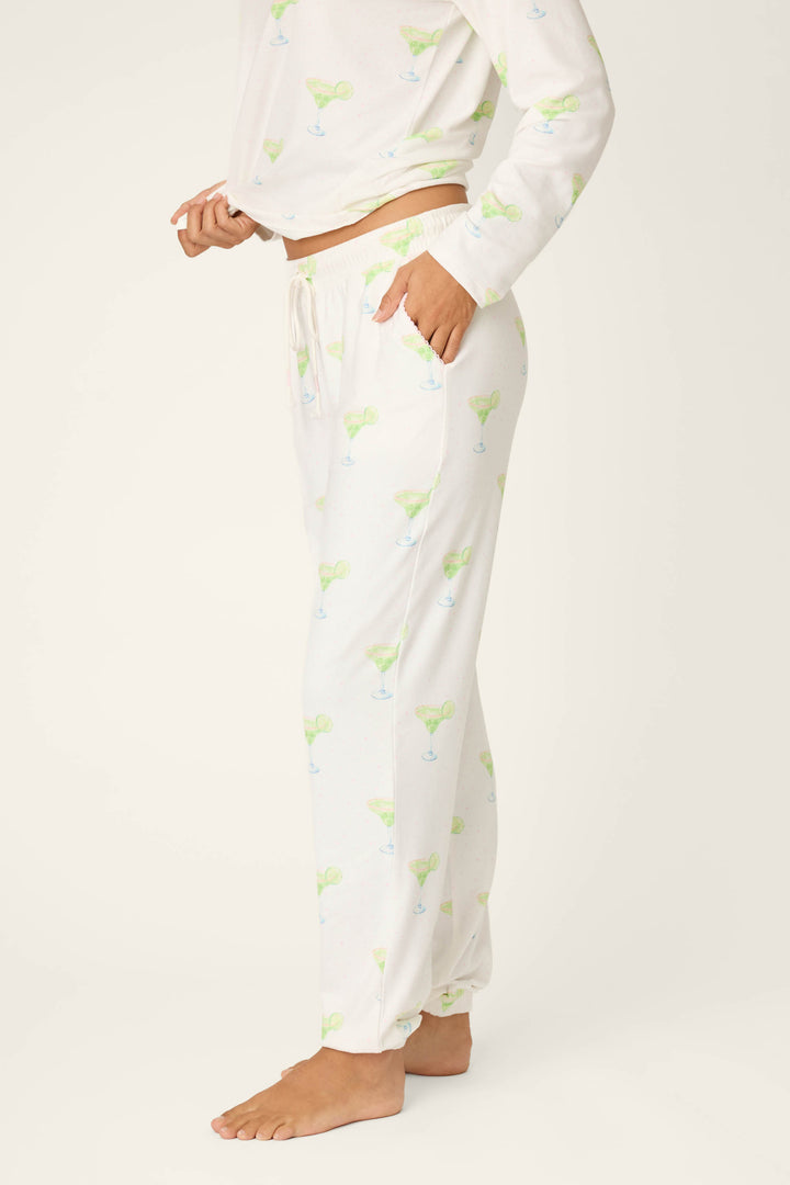 A person stands barefoot against a plain light background, wearing P.J. Salvage Margarita Mama Spicy Sips Banded Pants and a matching long-sleeve top in a green and blue margarita print made of buttery-soft jersey.