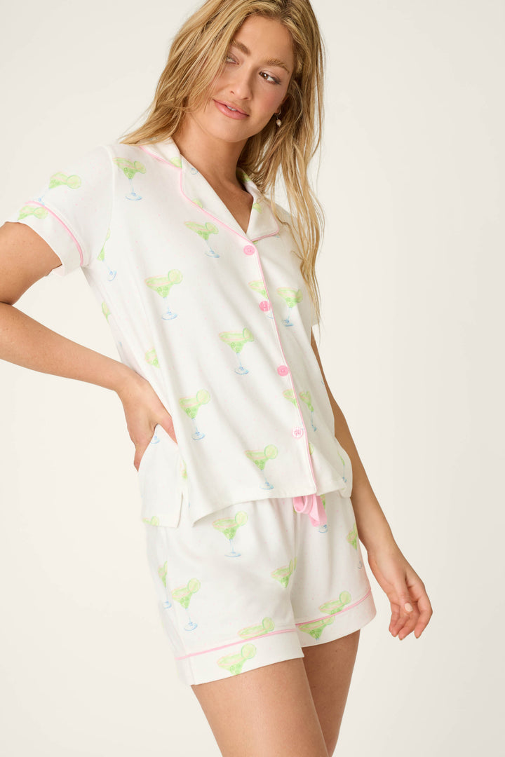 A woman with long blonde hair models the P.J. Salvage Margarita Mama Spicy Sips 3-Piece Short PJ Set, a collared, short-sleeve ensemble with pale margarita prints and pink accents, standing against an off-white background.