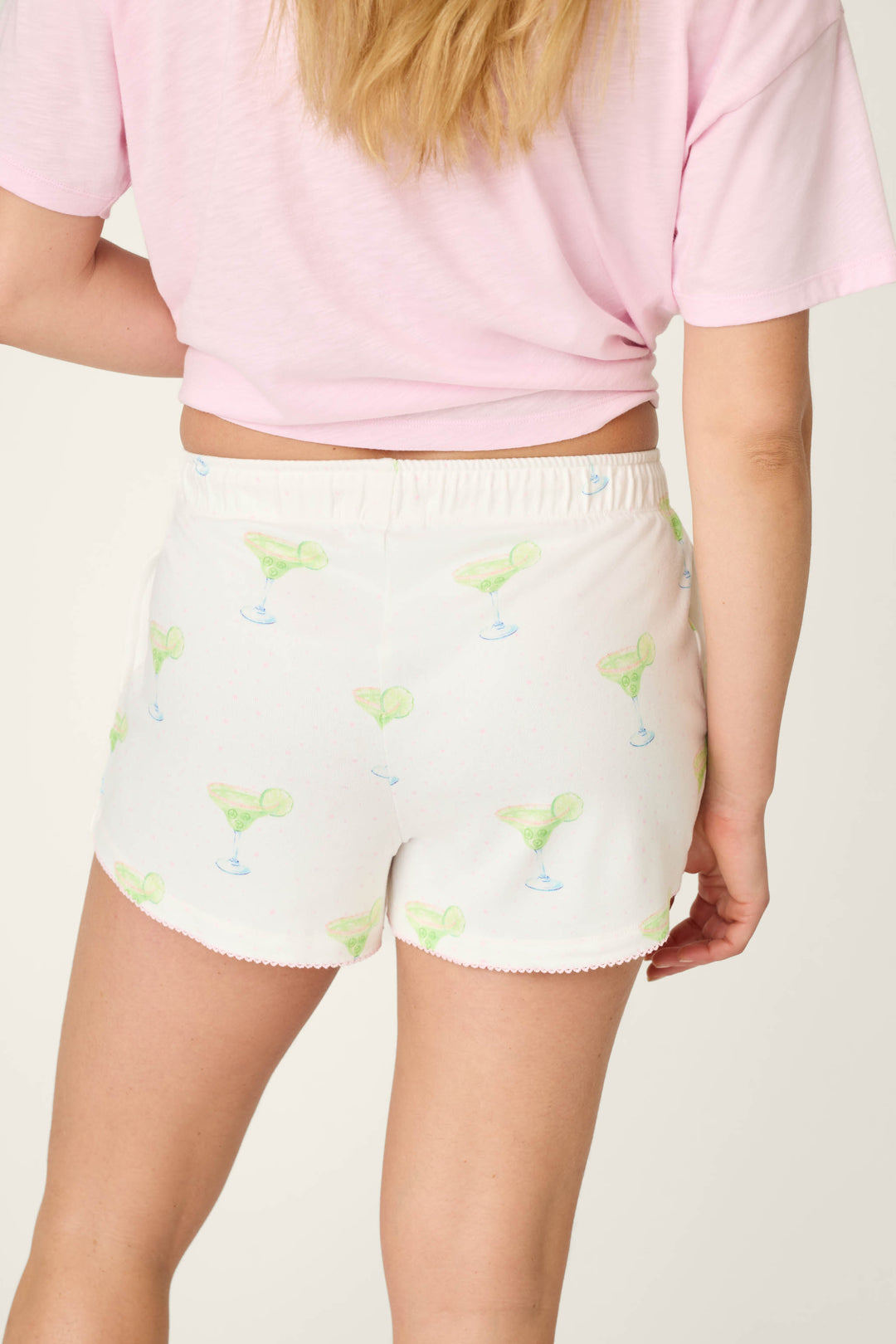 A person shown from the back wears a light pink T-shirt and P.J. Salvage Margarita Mama Spicy Sips Sleep Shorts—featuring buttery-soft jersey fabric, a green margarita print, elastic waistband, and scalloped hem.