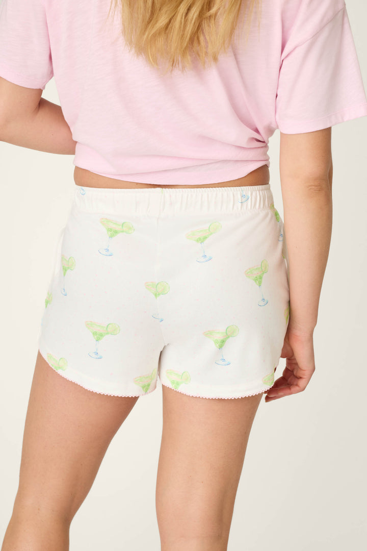 A person shown from the back wears a light pink T-shirt and P.J. Salvage Margarita Mama Spicy Sips Sleep Shorts—featuring buttery-soft jersey fabric, a green margarita print, elastic waistband, and scalloped hem.