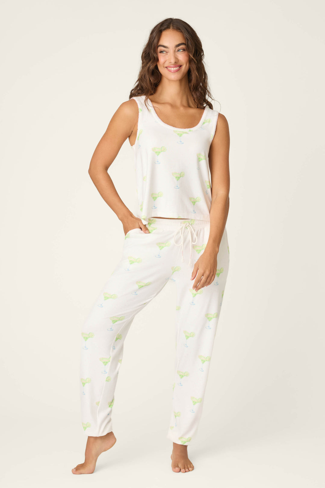 A woman stands barefoot, smiling, in P.J. Salvage's Margarita Mama Spicy Sips Tank Top—a sleeveless white pajama tank with a light margarita print and loose fit. The background is plain and light-colored, evoking relaxing vacation vibes.