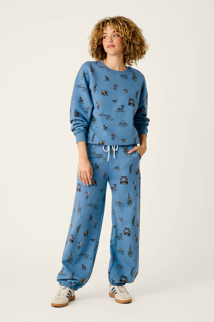 A person with curly hair wears the P.J. Salvage “Nature Is My Therapy” Nature Print Frankie Sweatshirt in blue, paired with matching sweatpants featuring small animal prints and a white drawstring, along with white sneakers with black stripes, standing against a plain light background.