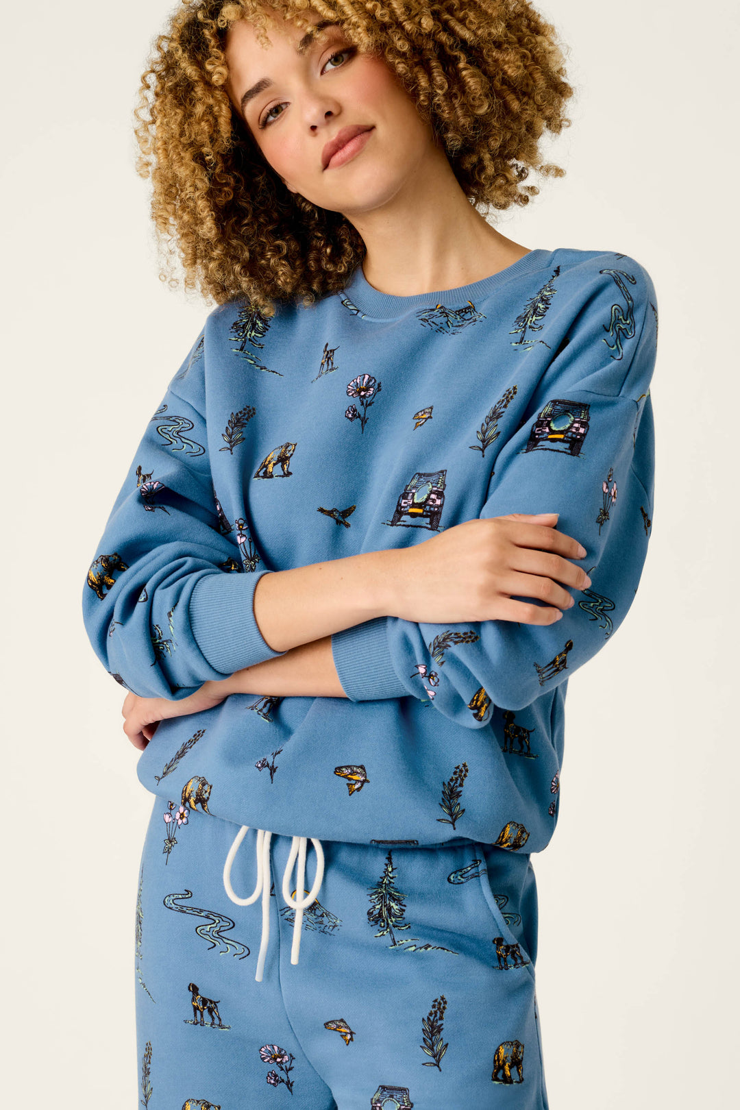 A woman with curly hair stands against a plain light background, arms folded and wearing the P.J. Salvage “Nature Is My Therapy” Nature Print Frankie Sweatshirt paired with matching sweatpants, both adorned with small nature and cabin illustrations. She smiles slightly.