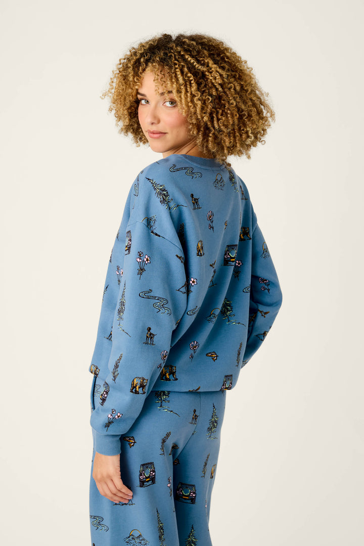 A person with curly hair wears the P.J. Salvage Nature Is My Therapy Nature Print Frankie Sweatshirt—featuring small outdoor-themed illustrations and paired matching pants—standing sideways and looking over their shoulder against a plain background.