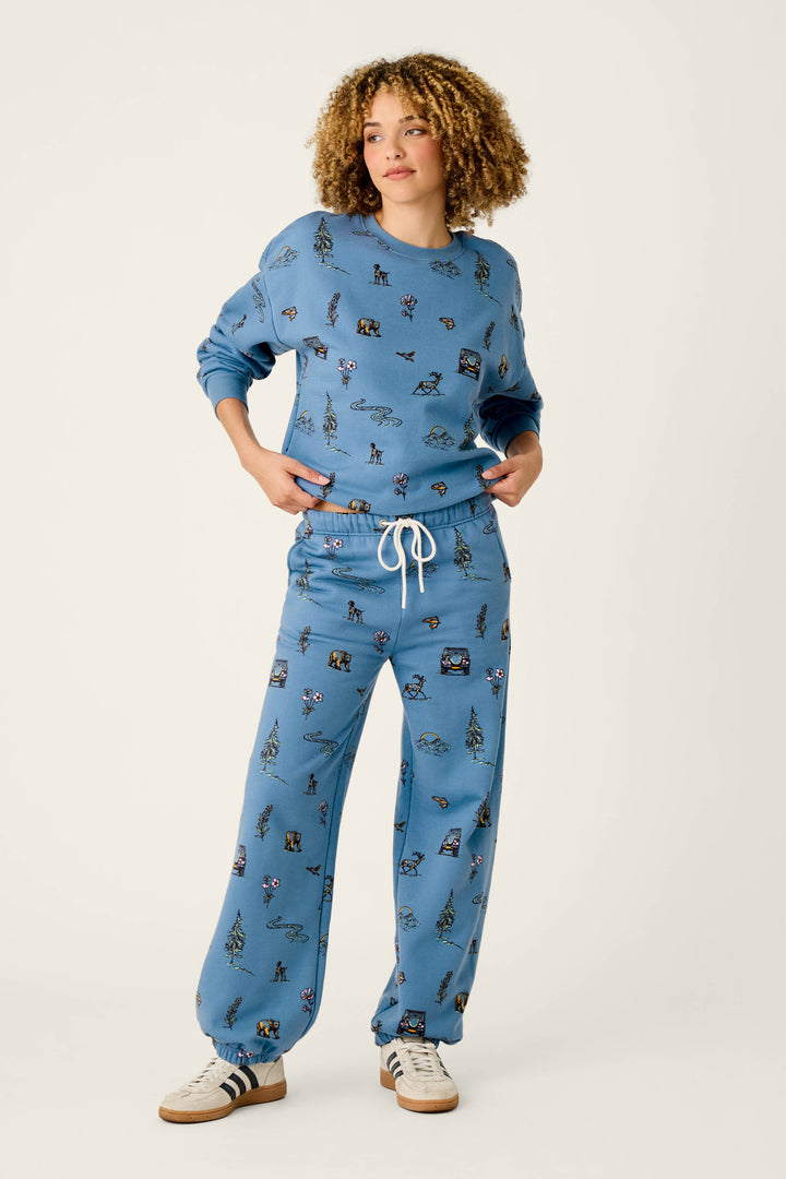 A person with curly hair wears the P.J. Salvage Nature Is My Therapy Nature Print Good Hang Joggers—a matching light blue sweatshirt and sweatpants set adorned with playful cartoon animal prints and a white drawstring—paired with white sneakers featuring dark stripes, standing against a plain background.