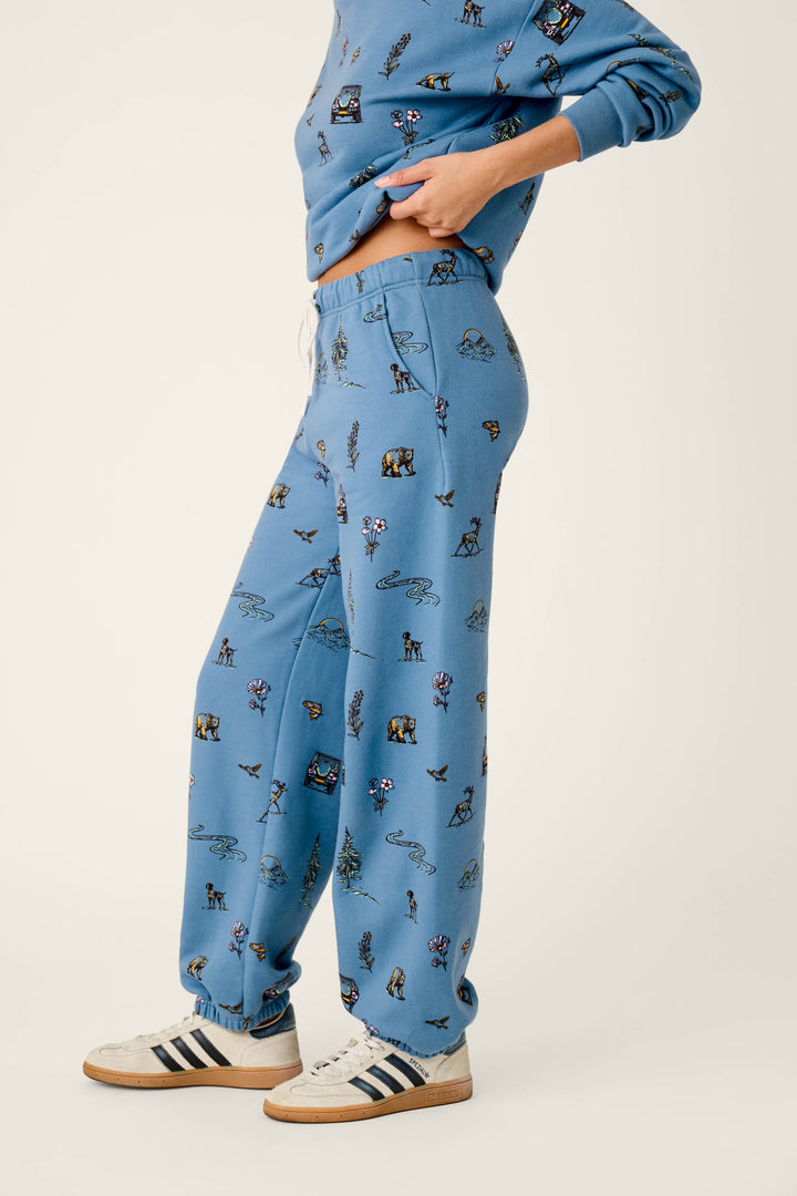 A person wearing the P.J. Salvage Nature Is My Therapy Nature Print Good Hang Joggers with a matching blue sweatshirt featuring small colorful prints, slightly lifting the sweatshirt to reveal their midriff, and white Adidas sneakers with navy stripes, posed against a plain light background.