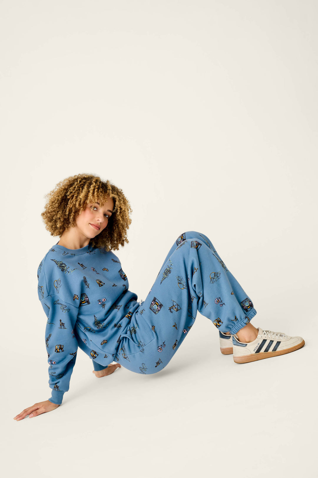 A person with curly hair wears a blue sweatshirt and P.J. Salvage Nature Is My Therapy Nature Print Good Hang Joggers, sitting on the floor and leaning back on one hand with legs bent and white Adidas sneakers.