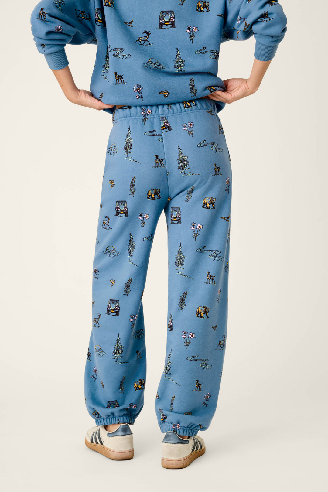 A person stands with hands on hips, wearing the P.J. Salvage Nature Is My Therapy Nature Print Good Hang Joggers—blue sweatpants featuring small outdoor-themed illustrations—paired with a matching sweatshirt. They have striped sneakers on, and the photo is taken from behind against a plain background.
