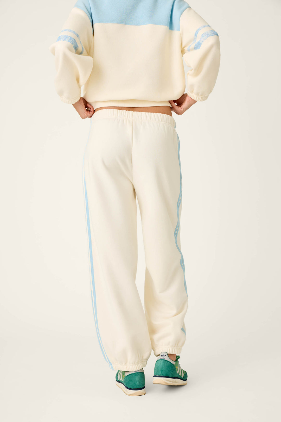 A person stands with their back to the camera, wearing the P.J. Salvage Nature Is My Therapy Adventure Awaits Good Hang Joggers—cream-colored sweatpants featuring light blue stripes down the sides—paired with a matching cream and blue sweatshirt and green sneakers.