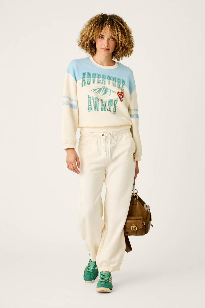 A woman with curly hair wears a cream and blue P.J. Salvage Nature Is My Therapy Adventure Awaits Margaux Sweatshirt, cream sweatpants, green sneakers, and carries a brown bag. She stands against a plain light background.