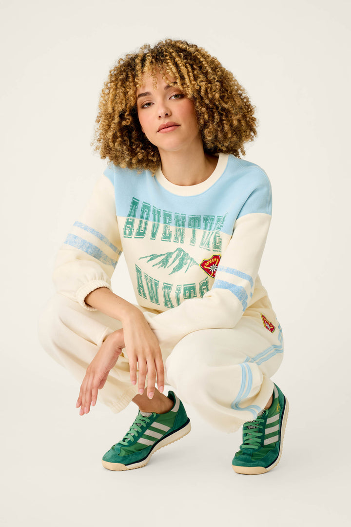 A woman with curly hair squats against a plain light background, wearing the P.J. Salvage Nature Is My Therapy Adventure Awaits Margaux Sweatshirt in light blue and cream, matching pants, and green Adidas sneakers.
