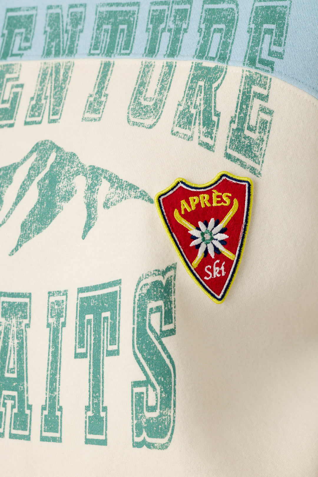 A close-up of the P.J. Salvage Nature Is My Therapy Adventure Awaits Margaux Sweatshirt featuring “AITS” in green, a mountain graphic, and a red shield patch reading “APRES Ski” with a white flower.