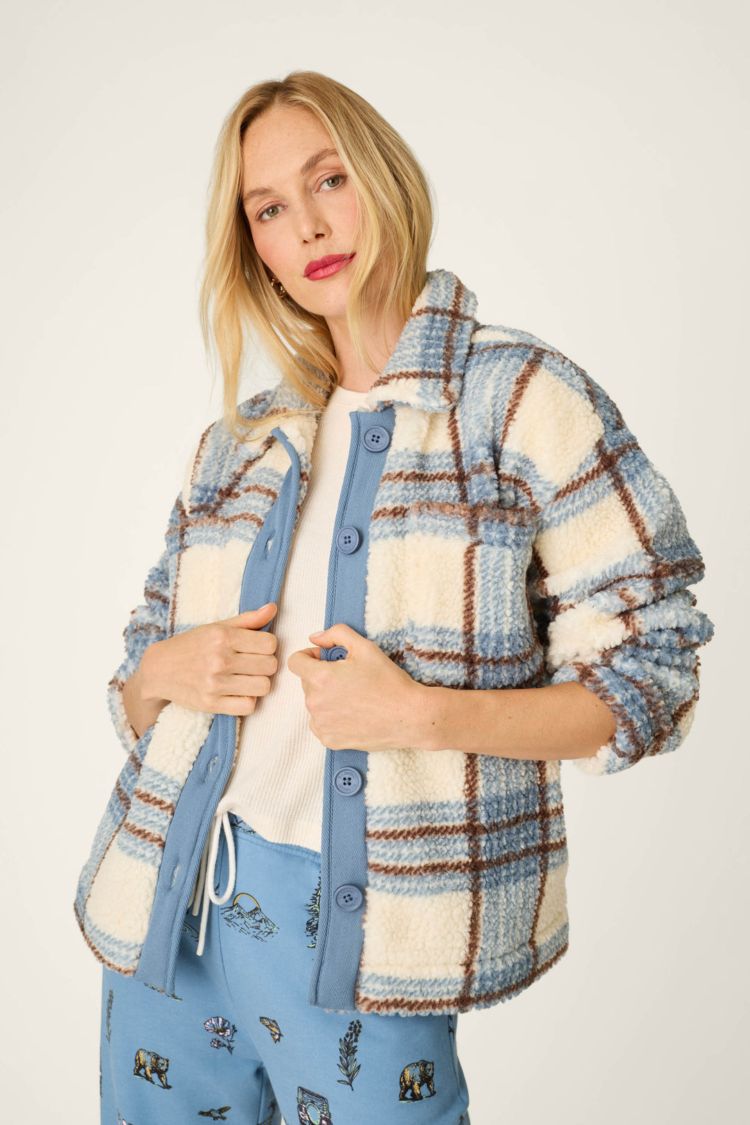 A woman wearing the P.J. Salvage Nature Is My Therapy Big Teddy Blue Plaid Sherpa Jacket over a white shirt and blue animal print pants, standing against a plain background.