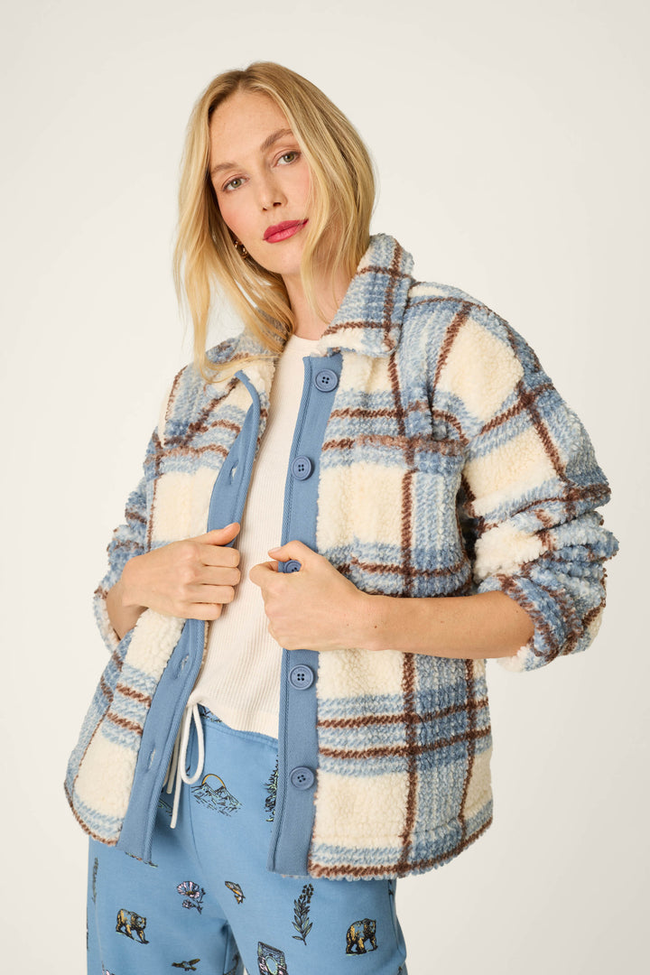 A woman wearing the P.J. Salvage Nature Is My Therapy Big Teddy Blue Plaid Sherpa Jacket over a white shirt and blue animal print pants, standing against a plain background.