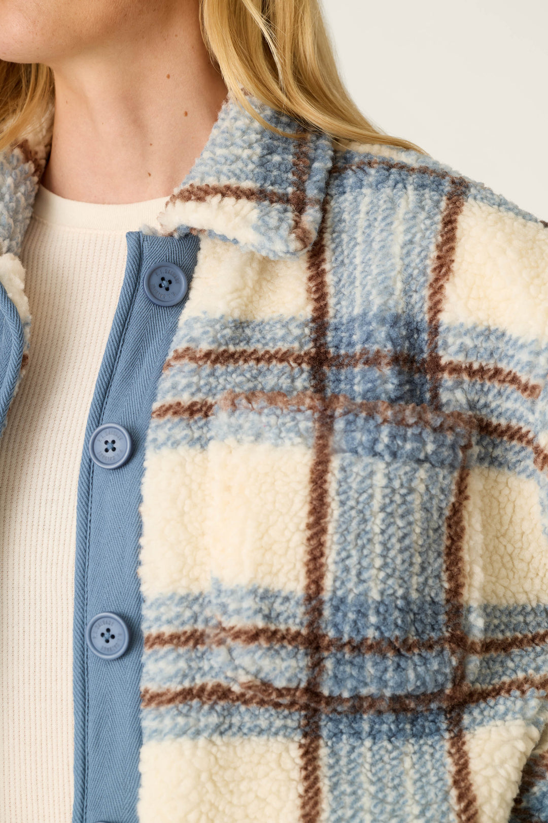 A close-up shows a person wearing the P.J. Salvage “Nature Is My Therapy Big Teddy Blue Plaid Sherpa Jacket”—featuring cream, blue, and brown plaid sherpa—layered over a ribbed white top and a blue buttoned shirt. Only their lower face and upper torso are visible.