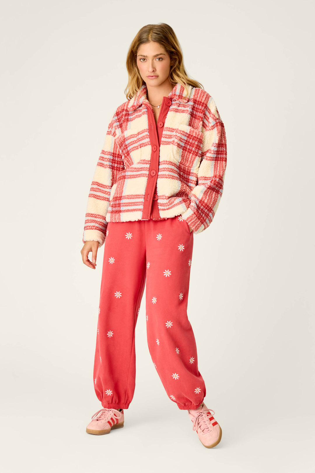 A woman with blonde hair stands against a plain, light-colored background wearing the Nature Is My Therapy Big Teddy Red Plaid Sherpa Jacket by P.J. Salvage over a red top and red pants with white daisy patterns. She completes her look with pink sneakers.