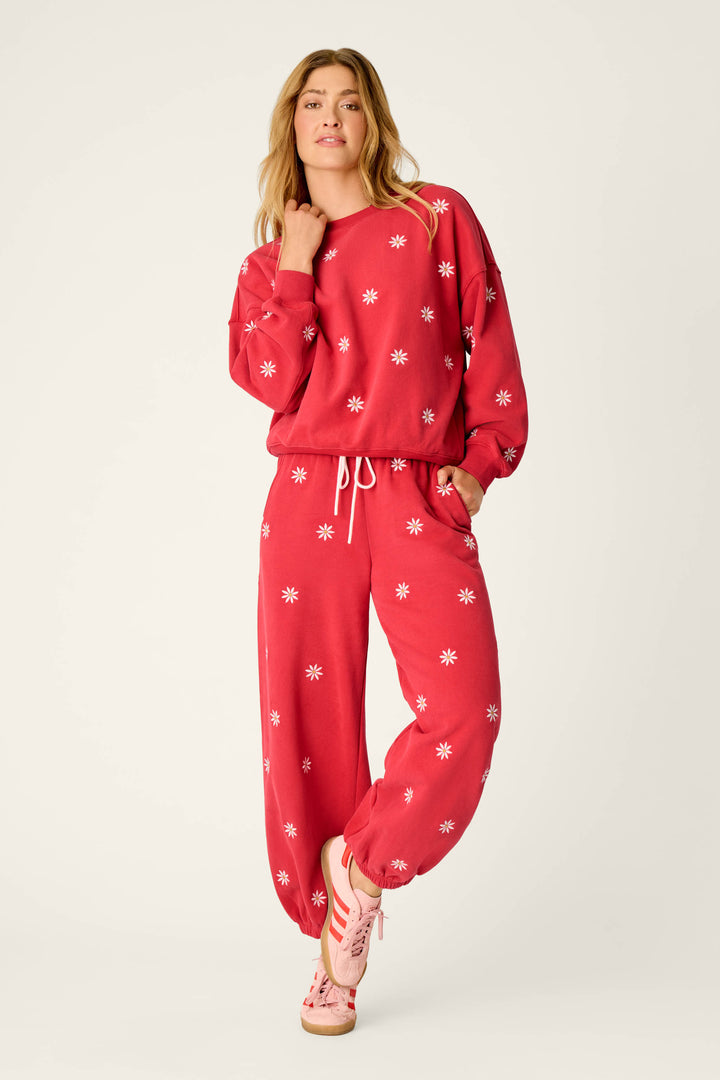 A woman wears P.J. Salvage’s Nature Is My Therapy Embroidered Flowers Good Hang Joggers, pairing them with a matching red sweatshirt featuring white starburst patterns and pink sandals, standing with one hand on her neck against a plain background.