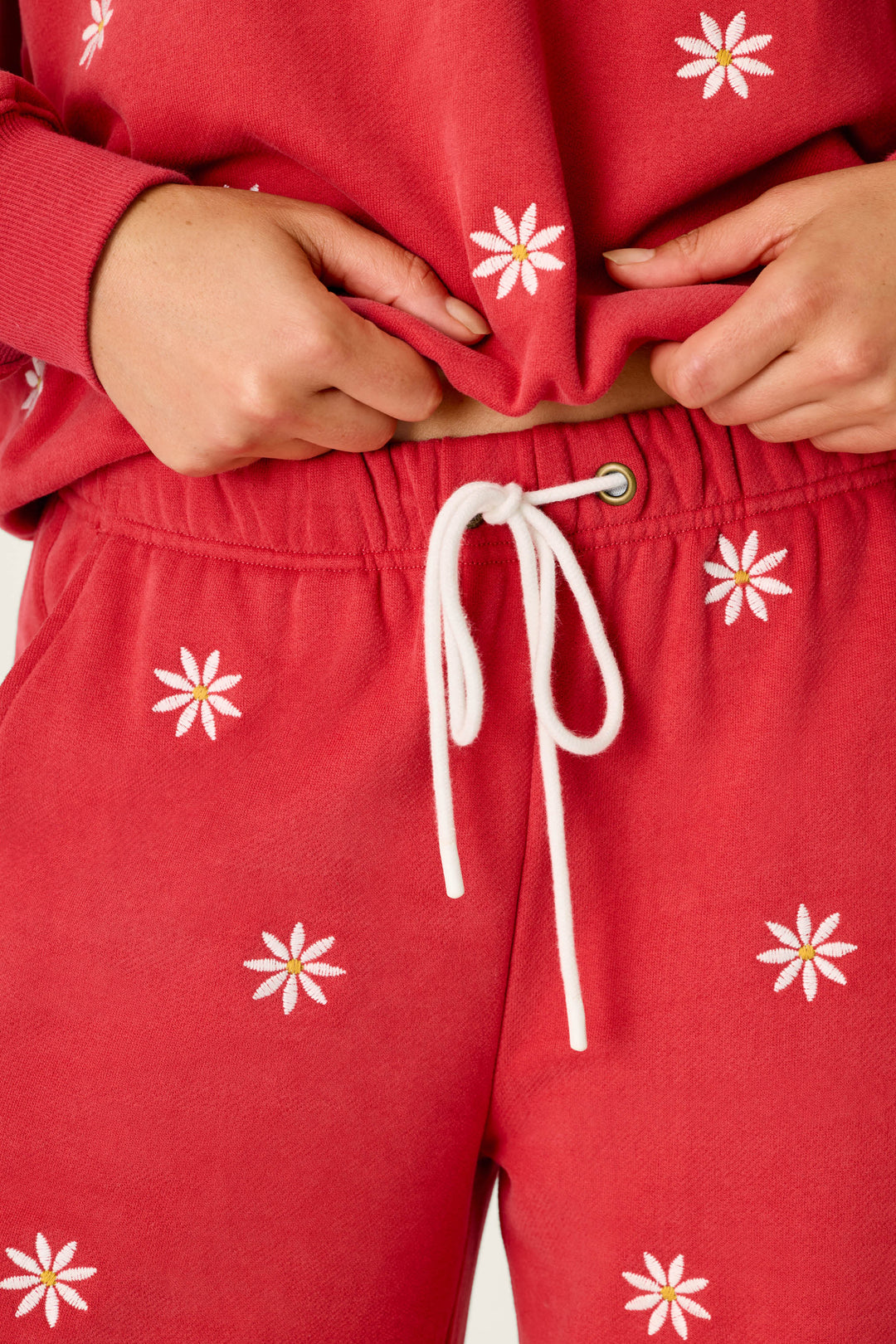 Wearing the P.J. Salvage Nature Is My Therapy Embroidered Flowers Good Hang Joggers, a person styles the matching red sweatshirt and joggers featuring white drawstrings and daisy embroidery, pulling up the top slightly to reveal the jogger’s waistband.